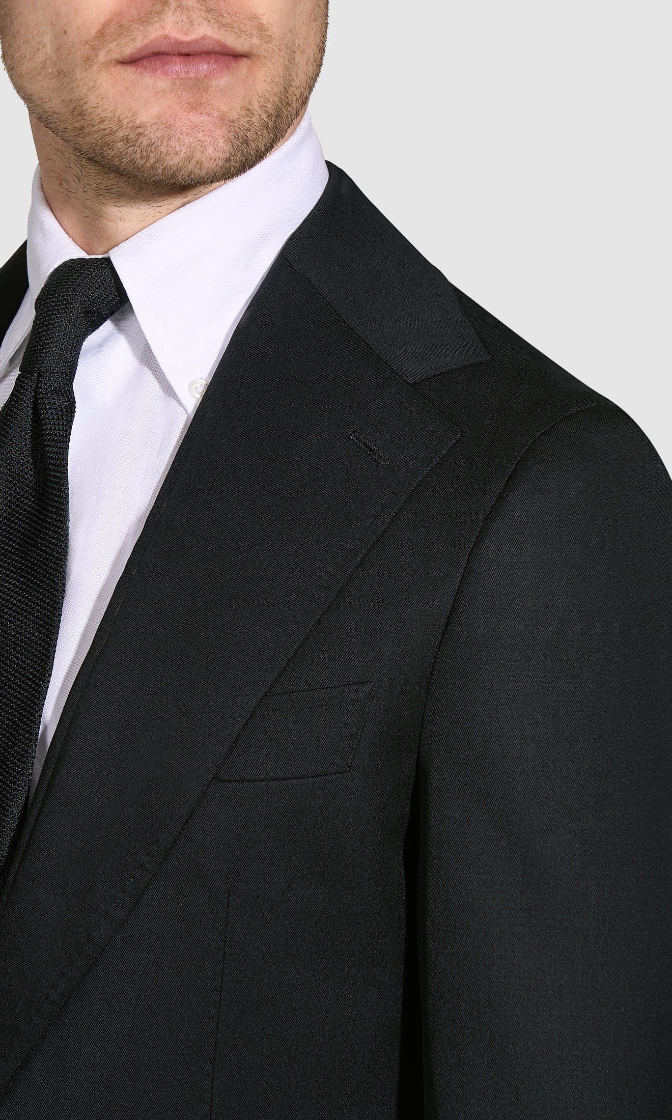 Man wearing a black suit jacket over a white shirt with a black tie on a light gray background