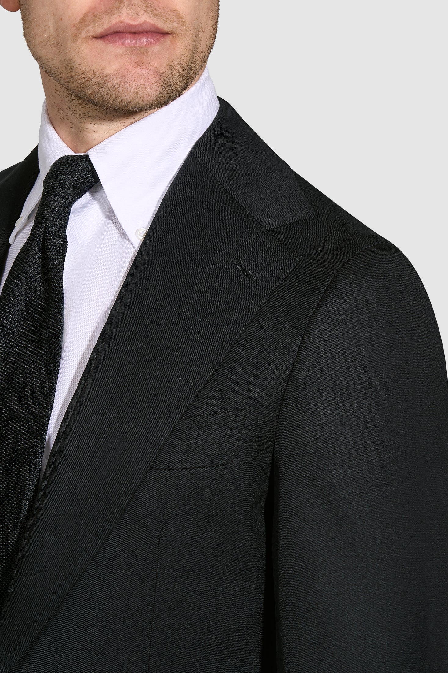 Man wearing a black suit jacket over a white shirt with a black tie on a light gray background